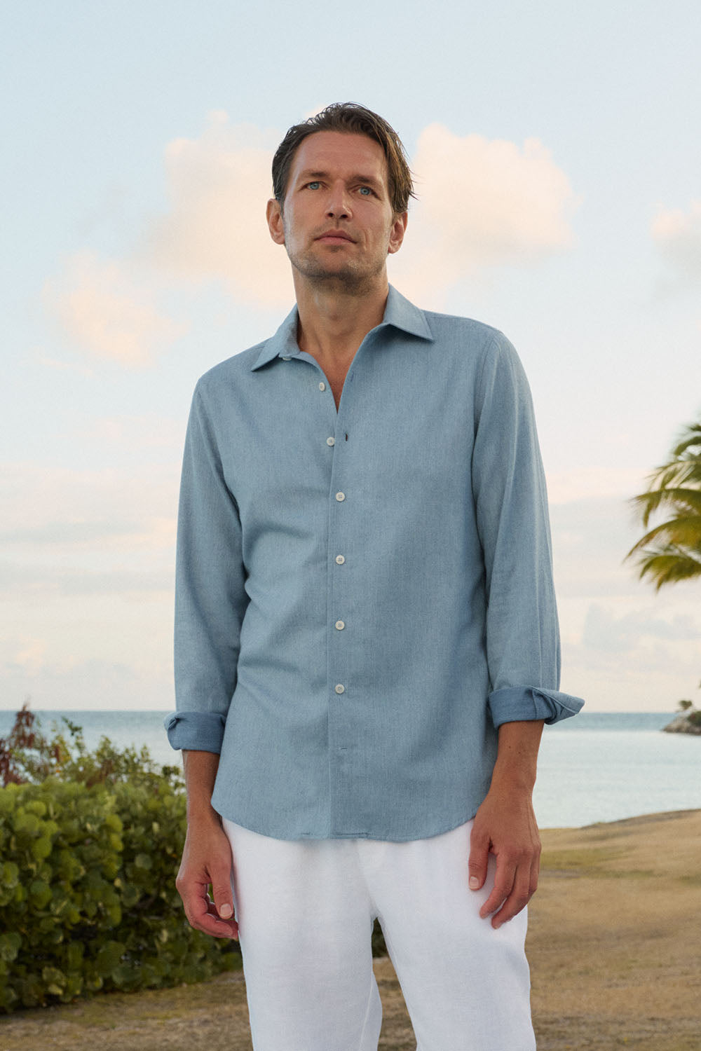 A man wearing a light blue long-sleeve linen button-up shirt and white pants, standing outdoors near a tropical beach with the ocean and sky in the background.