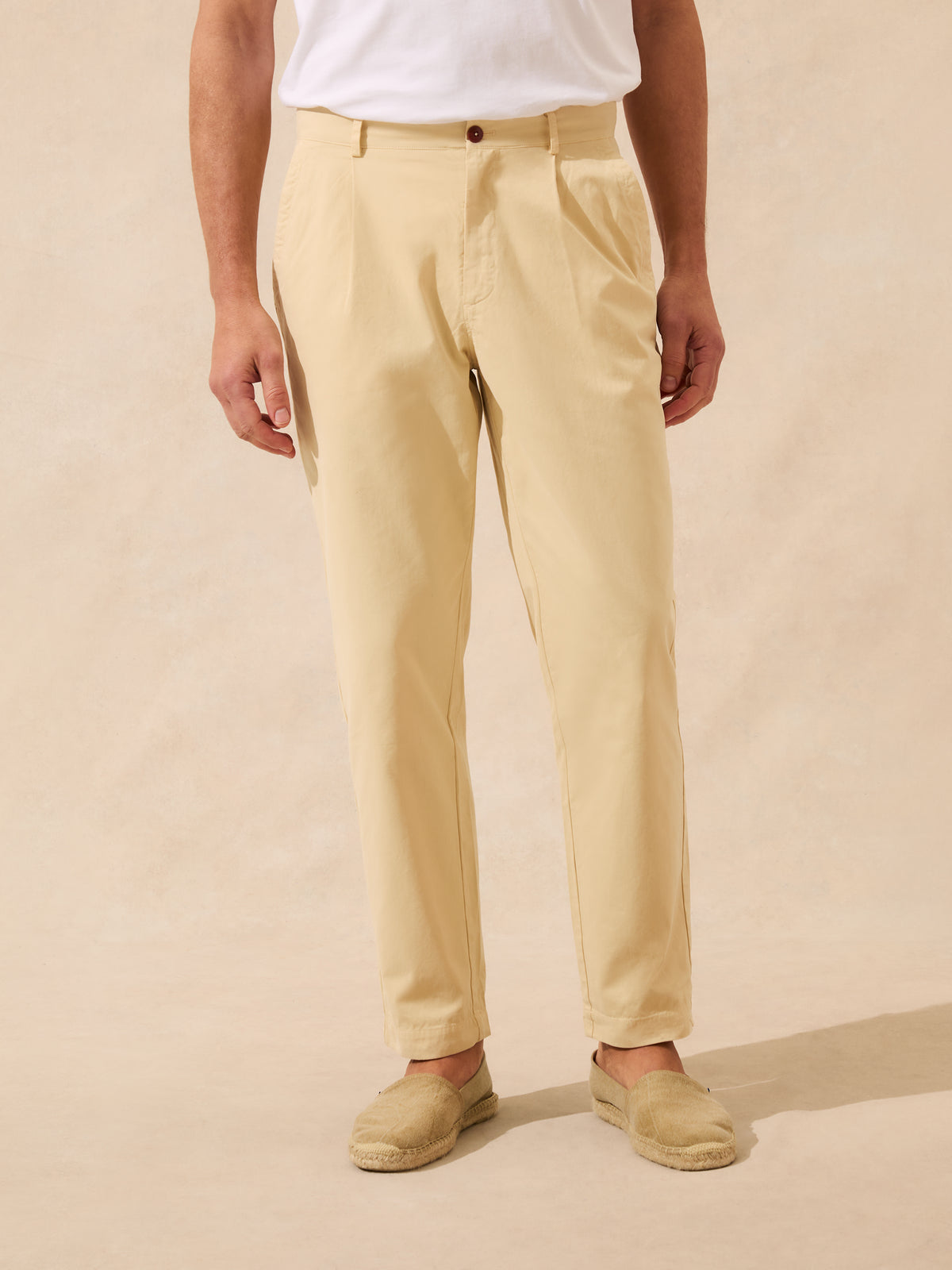 A person wearing light beige chinos with a white t-shirt and beige espadrilles, standing against a textured beige background.