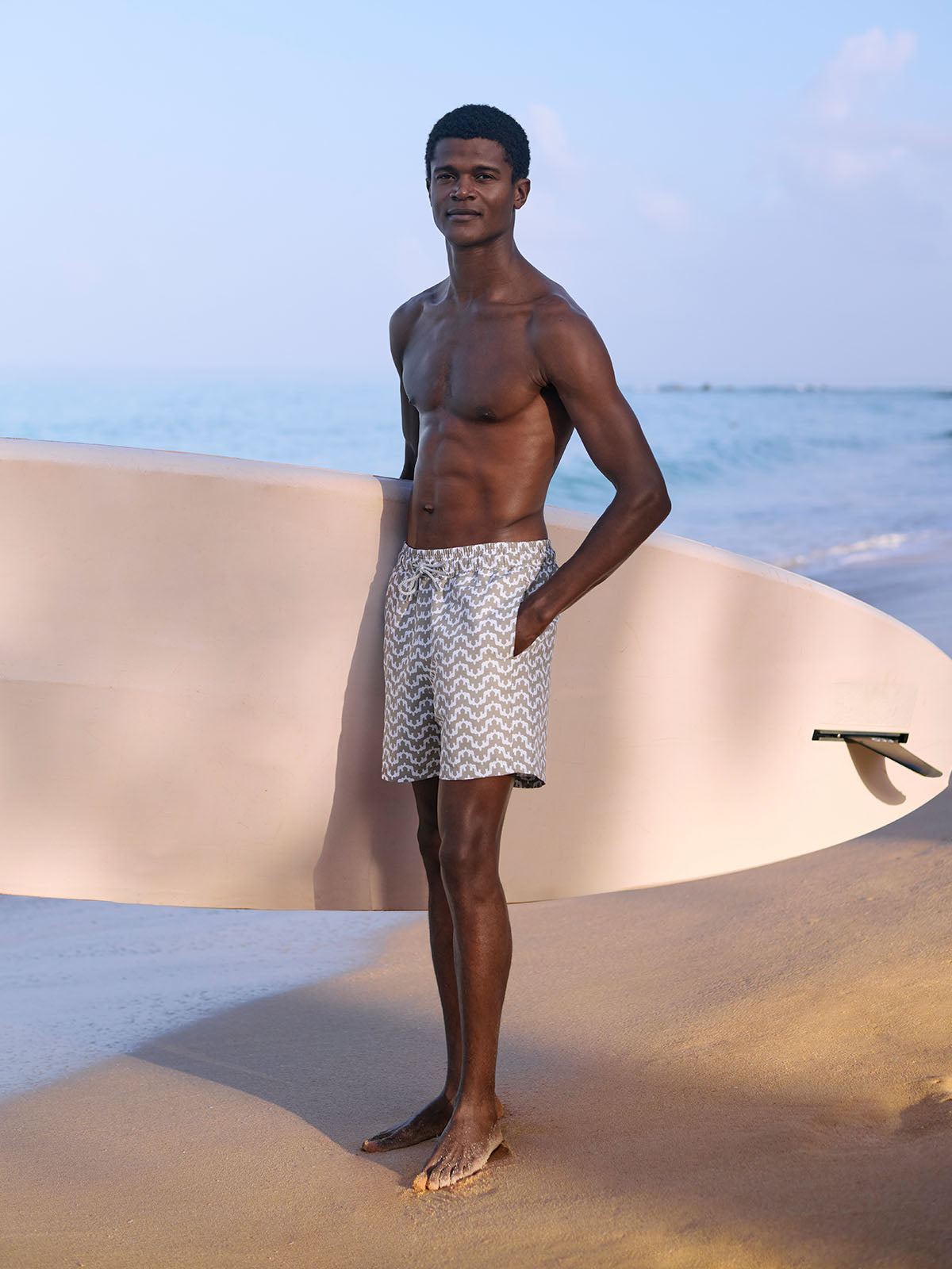 Man standing on a sandy beach holding a surfboard wearing green swim shorts