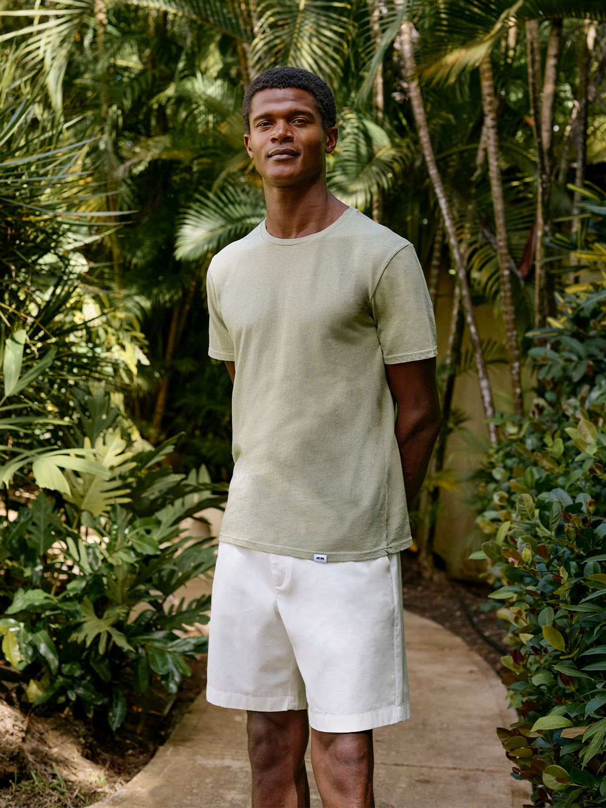 Man wearing a light green t-shirt and white linen shorts standing in a tropical setting with lush greenery.