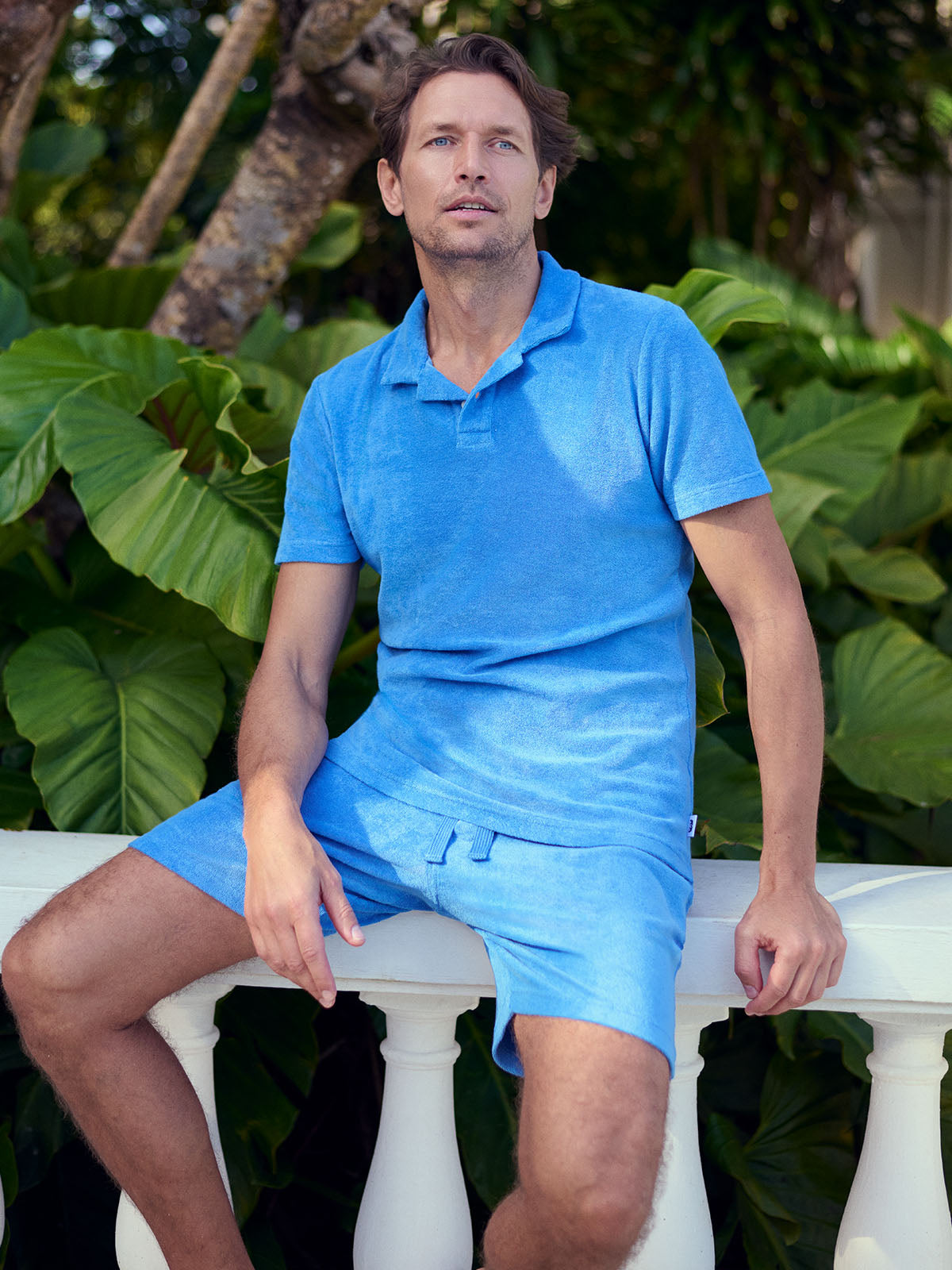 Man wearing a blue terry towelling outfit sitting on a white railing with green foliage in the background