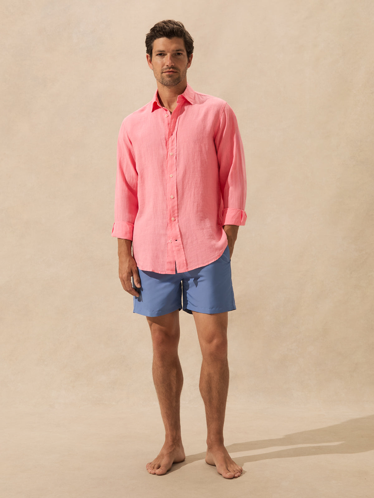 A man stands barefoot against a warm beige textured studio backdrop, wearing a pink long-sleeve button-up shirt and blue swim shorts.