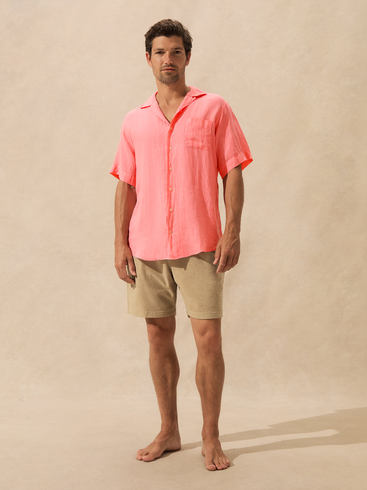 A man stands barefoot against a warm beige studio backdrop, wearing a short-sleeve coral-pink button-up shirt and light khaki shorts.