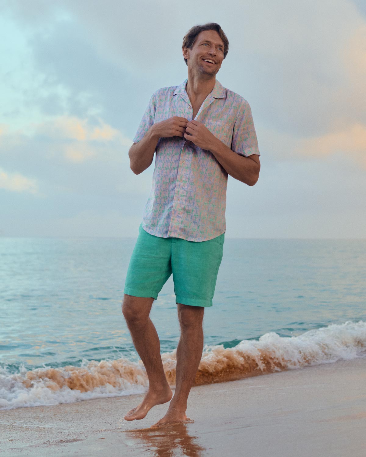 Man standing on a beach wearing a light pink shirt and green shorts with a sunset in the background.