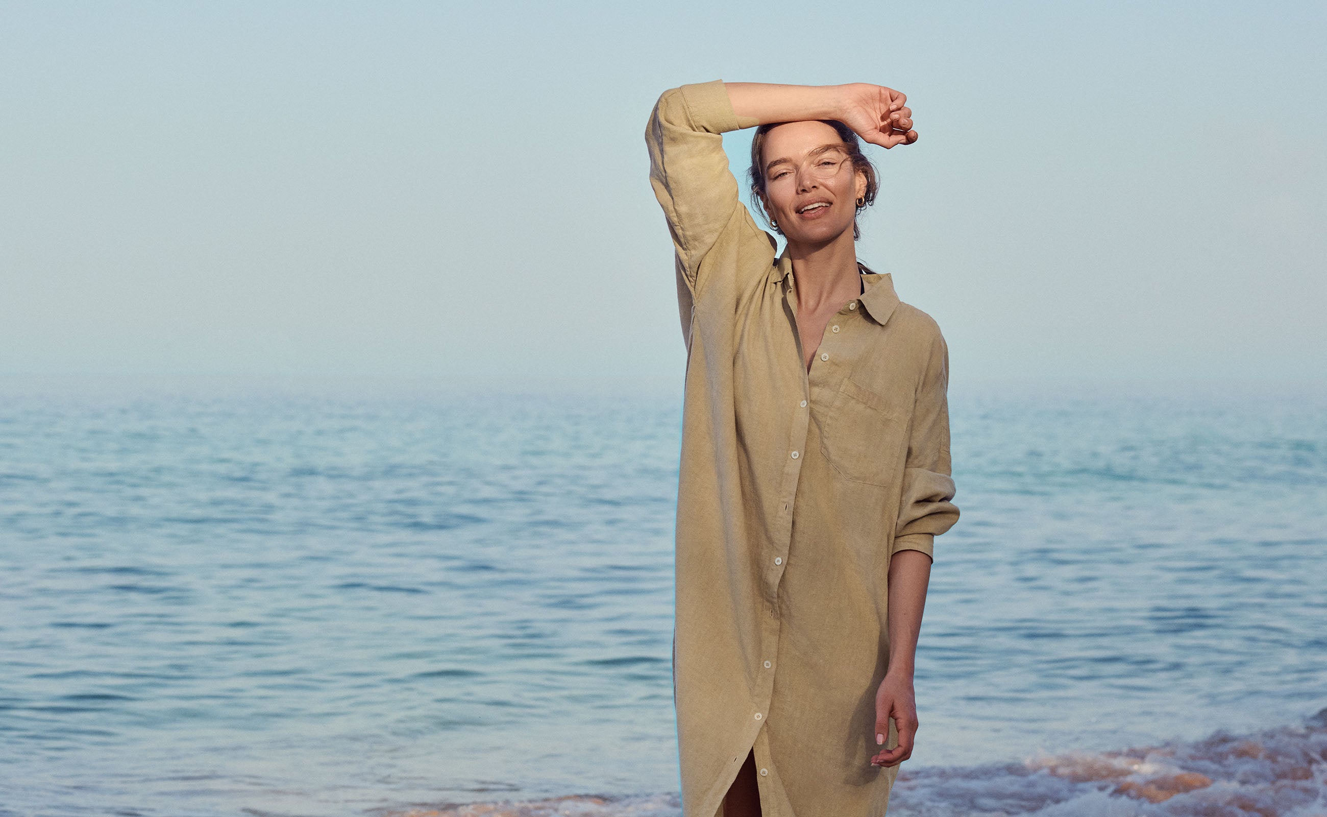 Woman in a beige dress by resort wear uk brand standing on a beach with ocean in the background
