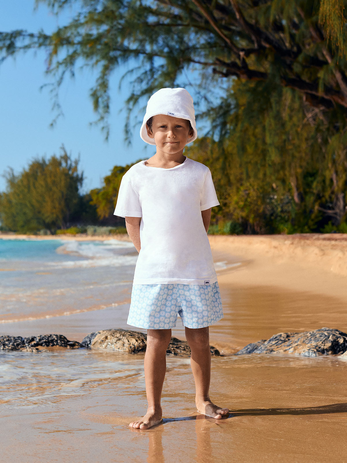Child standing on a beach wearing a white shirt and patterned swim shorts with a hat.