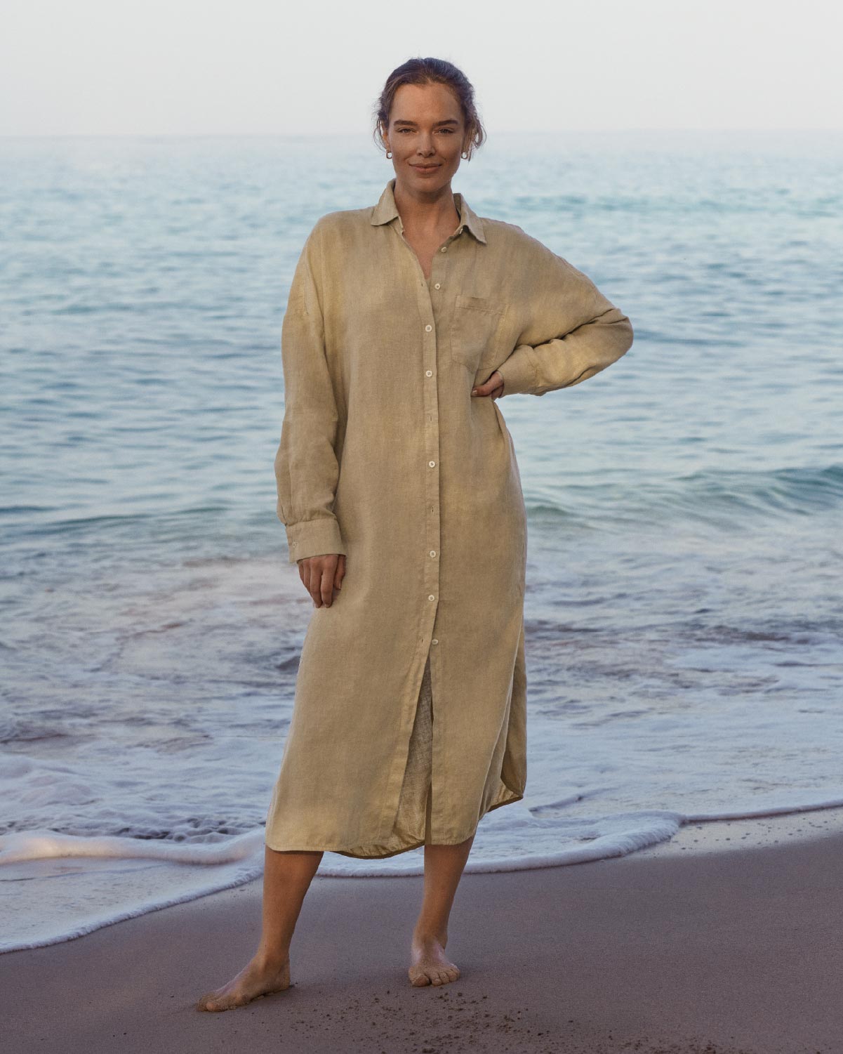 Woman wearing a beige dress standing on a beach with ocean waves in the background