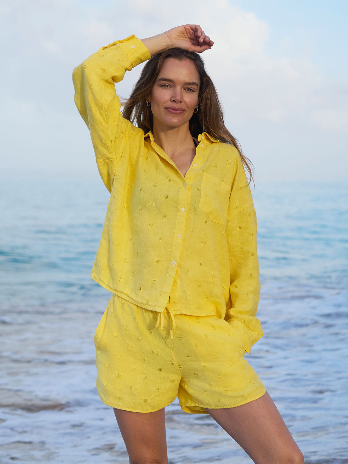 Woman in a yellow linen shirt and linen shorts standing on a beach with ocean in the background