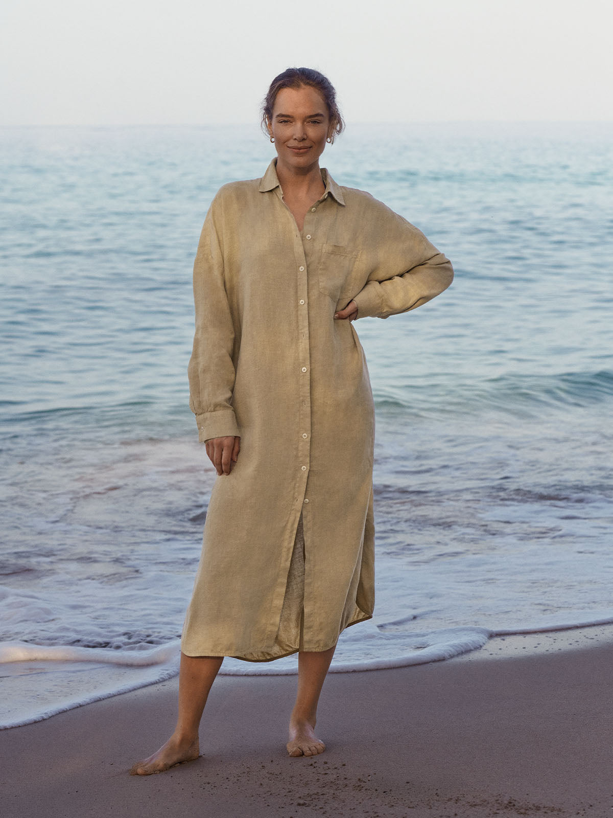 Woman in a beige dress by resort wear uk brand standing on a beach with ocean waves in the background